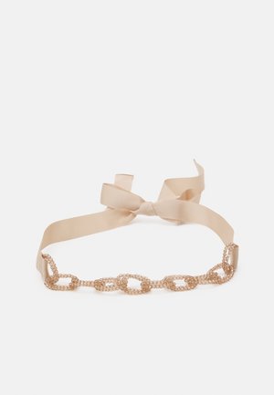 Collier ras-du-cou beige en ruban avec ornements de strass ovales liés et fermeture à nœud papillon sur fond blanc.