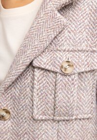 Tekstureeritud, helesinine herringbone mantli, millel on krae ja suur nuppe detail. Omab rinna taskut klapiga.