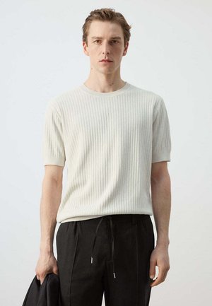 Joven con un suéter blanco de manga corta con ribetes y pantalones negros con cordón, sosteniendo una chaqueta negra en su mano izquierda.