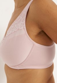 Frau trägt einen hellrosa BH mit Spitzenapplikation auf den Körbchen, gezeigt aus der Nahaufnahme seitlich mit Fokus auf Passform und Stoffstruktur.