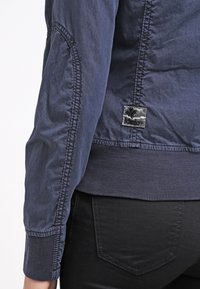 Gros plan d'une personne portant une veste bleue avec des poignets et un ourlet côtelés, et un pantalon noir avec une poche arrière visible.