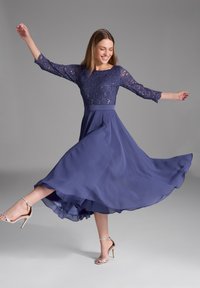 Robe longue navy avec corsage en dentelle, manches trois-quarts et jupe en chiffon fluide. Associée à des sandales à talons hauts argentées.