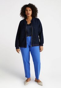 Mörkblå texturerad bomberjacka med dragkedja, tillsammans med en blå satinblus och skräddarsydda blå byxor. Vita skor utan snörning med detalj.