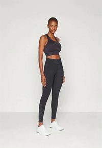 Fekete sportmelltartó, amely crop dizájnnal rendelkezik, fekete magas derekú leggingssel párosítva, amely hálós részletekkel és sima textúrával büszkélkedhet. Fehér cipők.