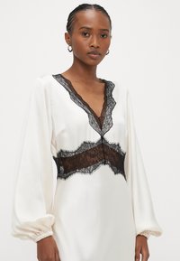 Balta satīna kleita ar garām piedurknēm, dziļu V-veida izgriezumu un melna mežģīņu akcents pie kakla un vidukļa, ar strukturētām pūderveida piedurknēm.