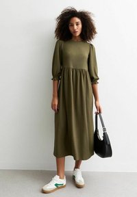 Robe midi vert olive avec des manches bouffantes, en tissu texturé, détails volants à la taille, associée à des baskets blanches et un sac à main noir.
