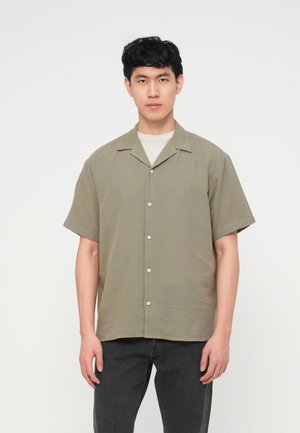 Νεαρός άντρας με σκούρα κατσαρά μαλλιά φοράει ελαφρύ χακί πουκάμισο με κοντά μανίκια και σκούρο τζιν με φόντο ένα απλό λευκό φόντο.