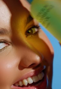 Närbild av ett ansikte med slät hy och ett lysande leende. Ett öga har en gyllene glans, medan en grön flaska delvis är synlig i förgrunden.