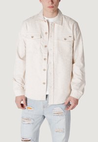 Giacca button-up beige chiaro con due tasche sul petto, un sottile motivo testurizzato e un colletto classico, abbinata a jeans leggeri blu destrutturati.