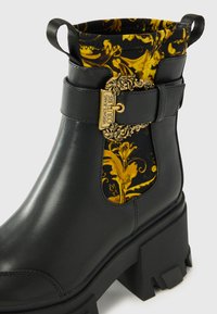 Black leather ankle boot with a chunky heel, patterned yellow and black lining, decorative gold buckle, and pull tabs on the collar.