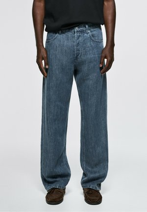 Homme portant un jean bleu ample, des chaussures marron foncé et une chemise noire sur un fond gris uni.