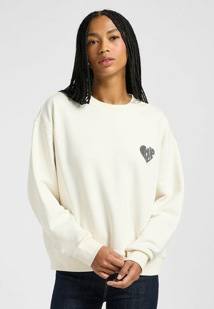 Sudadera de color crema con un corte relajado, que presenta un diseño de corazón en el lado izquierdo del pecho. Tiene puños y bajo acanalados.
