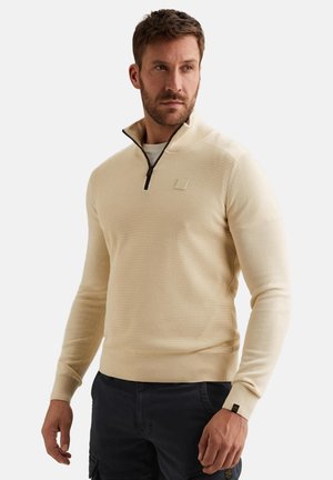 Mężczyzna w beżowym swetrze z półzamkiem i teksturowanym przodem oraz ciemnych spodniach, patrzący w prawo na tle jednolitego, jasnego tła.