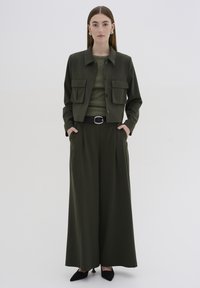 Olivengrüne, verkürzte Jacke mit Fronttaschen, kombiniert mit passenden weit geschnittenen Hosen und einem figurnahen Oberteil. Ein schwarzer Gürtel und spitze Schuhe runden das Outfit ab.