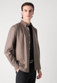 Barna csíkozott, galléros ing, hosszú ujjakkal és fehér gombokkal, egy fekete ing felett viselve, fekete nadrággal párosítva.