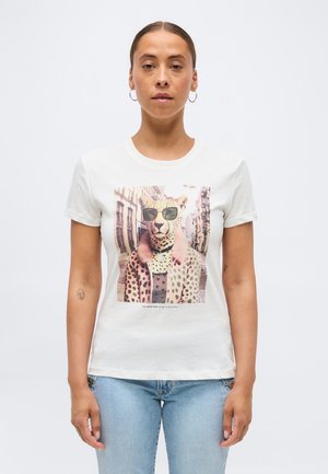 Frau trägt ein weißes T-Shirt mit einem gedruckten Motiv von einem Geparden mit Sonnenbrille und Pelzmantel, steht vor einem schlichten Hintergrund.