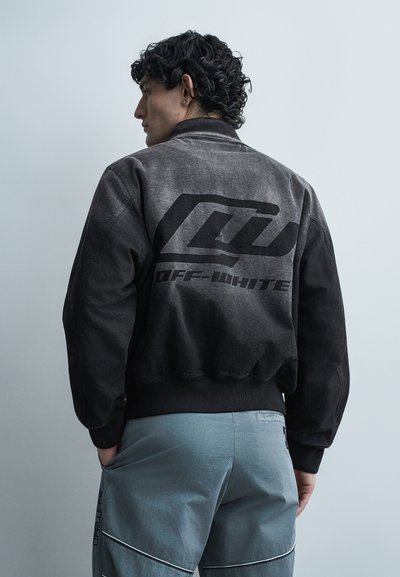 Homme aux cheveux noirs bouclés portant une veste Off-White noire délavée de profil et un pantalon bleu avec des poches.