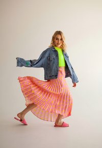 Giubbotto di denim con dettagli ricamati, top corto neon verde, gonna a pieghe rosa con pois gialli e sandali rosa senza lacci.