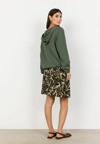 Olívovo zelená mikina s kapucňou v kombinácii so sukňou so vzorom palmových listov v zemitých farbách. Hnedé ploché topánky dotvárajú celý outfit.