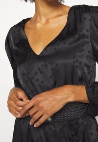 Černá saténová blůza s výstřihem do V, s texturovaným puntíkovým vzorem a nařasenými rukávy, doplněná tkaným černým páskem.
