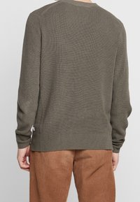Olivgrüner gerippter Pullover mit langen Ärmeln, Rundhalsausschnitt und seitlichen Etiketten. Das Material hat eine weiche Textur. Kombiniert mit Cordhosen.