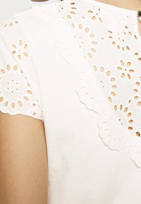 Biały bawełniany top z misternym haftowanym przetykaniem przy kołnierzu. Krótkie rękawy i okrągły dekolt. Gładka tekstura.