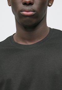 Tmavě zelené triko s kulatým výstřihem vyrobené z hladké bavlněné tkaniny, s klasickým designem a švy, které zajišťují pohodlné nošení.