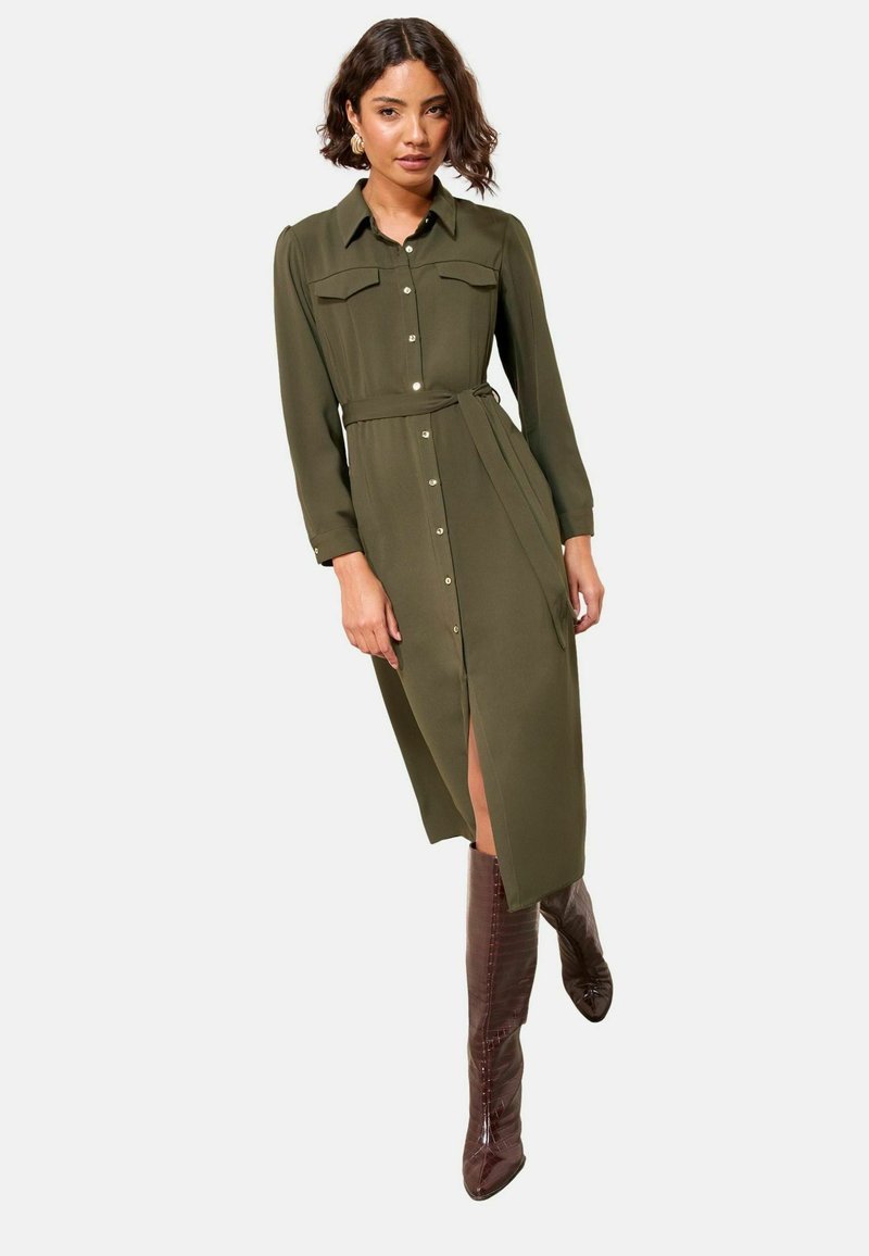 Oliven grøn, langærmet skjortekjole med knaplukning, to brystlommer, bælte i taljen og en slidse til låret. Parret med skinnende brune støvler.