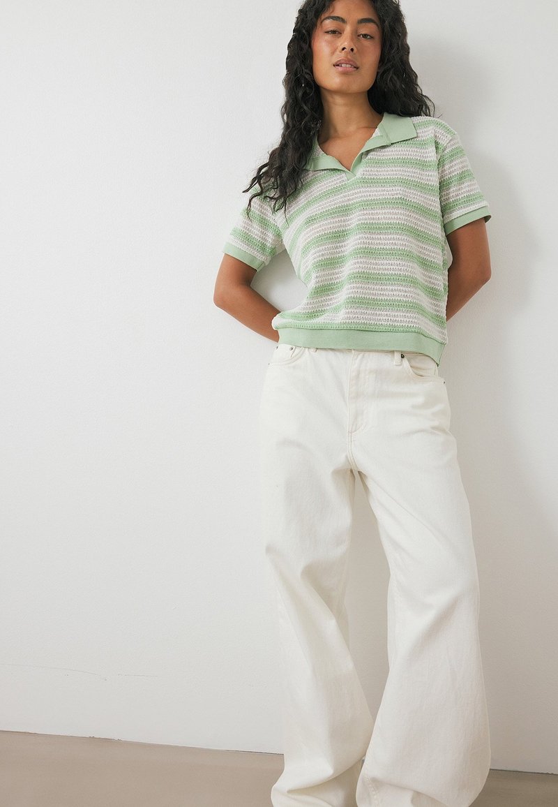 Sieviete ar garām, cirtainām matiem, tērpusies zaļi baltā svītrainā īsroku džemperī un baltās platajās biksēs, atbalstoties pie baltas sienas.