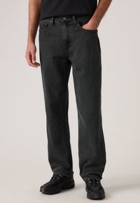 Μαύρο τζιν με ρίγες, χαλαρή εφαρμογή, με πέντε τσέπες, κλείσιμο με κουμπί και συνδυασμένο με μαύρα αθλητικά παπούτσια.