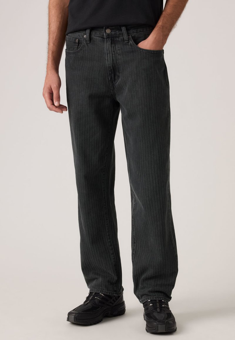 Jean en denim rayé noir avec une coupe décontractée, doté de cinq poches, fermeture par bouton, porté avec des baskets noires.