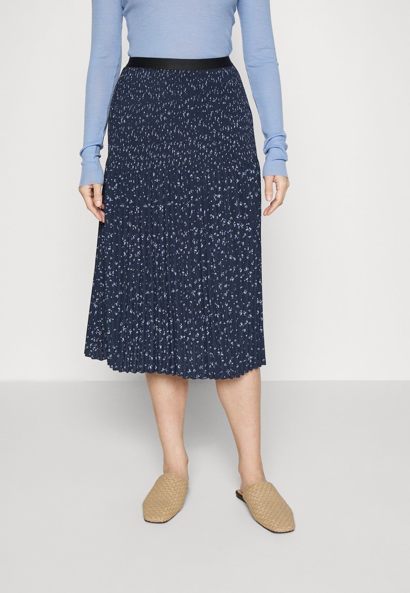 Námořnická plisovaná sukně s malými bílými květinovými vzory, vybavená černým pasem. Nosí se s tkanými béžovými nazouvacími botami.