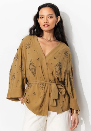 Femme portant un chemisier cache-cœur moutarde orné de perles noires, manches larges, ceinture nouée, pantalon blanc et bijoux en or.