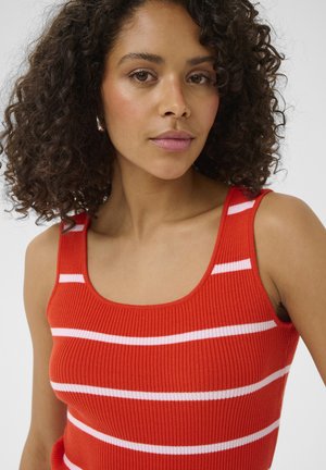Woman with curly dark hair wearing a red sleeveless ribbed top with white horizontal stripes, facing forward against a plain background.