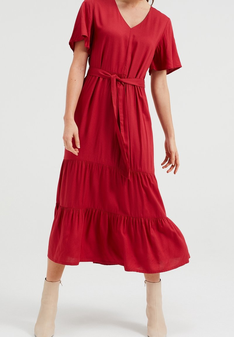 Červené midi šaty s krátkými volánkovými rukávy, výstřihem do V a přiléhavým páskem v pase. Mají stupňovité volány na lemu a jsou vyrobeny z lehké látky.