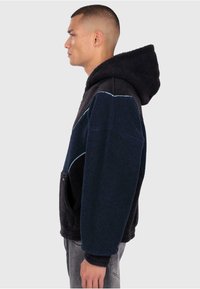 Fleecehuva med en marinblå och svart färgschema, som har en framficka, en huva och kontrasterande ljusblå sömmar. Strukturerad tyg.