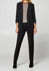 Svart skräddarsydd kavaj över en mönstrad topp, kombinerat med slim fit svarta byxor och svarta ankelstövlar, vilket skapar en elegant, monokromatisk look.