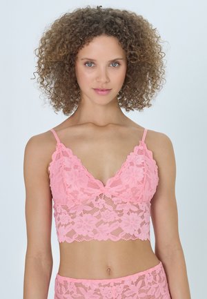 Młoda kobieta z kręconymi włosami, ubrana w różowy koronkowy biustonosz typu bralette w kwiaty i dopasowane majtki, stoi na jasnym, jednolitym tle.