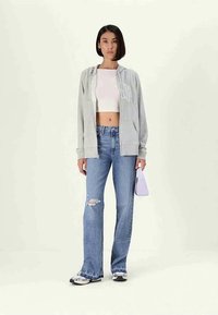 Mulher vestindo um hoodie cinza com fecho éclair, uma camisola curta branca, jeans azuis rasgados, ténis e segurando uma pequena bolsa lavanda.