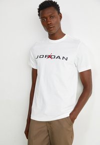 Camiseta blanca de algodón con un llamativo logo negro de "JORDAN", acentuado por un gráfico rojo, combinado con pantalones cortos beige. Diseño limpio y minimalista.