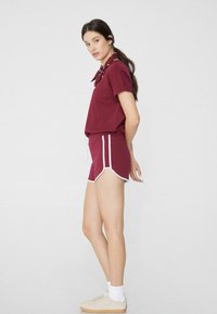 Haut bourgogne à manches courtes avec un col, associé à un short bourgogne assorti avec bordures blanches. Le modèle porte des baskets légères et des chaussettes blanches.