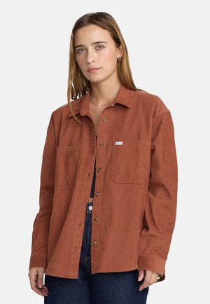 Jeune femme aux cheveux châtain clair porte une chemise en velours côtelé couleur rouille sur un haut noir et un jean bleu.