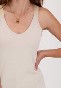 Vrouw in een beige mouwloos top met gouden bandjes, hand op de heup, en een gouden ketting met een klein hanger.