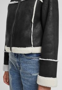 Veste en shearling noir avec des accents en polaire blanche, dotée d'une fermeture éclair, d'un design court et d'une texture contrastante le long des ourlets et des poignets.