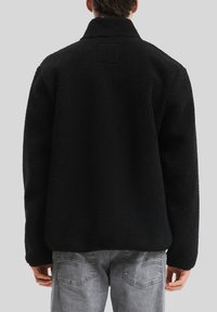 Schwarze Fleecejacke mit strukturierter Oberfläche, hohem Kragen und einem quadratischen Aufnäher auf dem Rücken. Über grauen Jeans getragen.