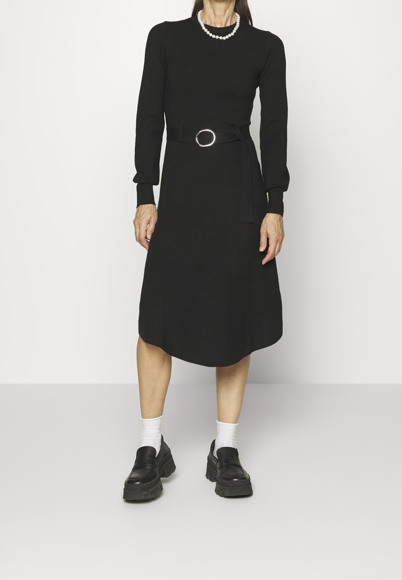 Femme portant une robe midi noire à manches longues avec ceinture, des chaussettes blanches, des mocassins épais noirs et un collier de perles, debout sur un sol clair.