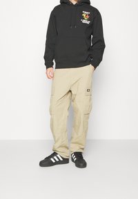 Černá mikina s kapucí a grafickým potiskem, béžové cargo kalhoty s kapsami a černé tenisky s bílými pruhy. Vzdušný streetwear styl.