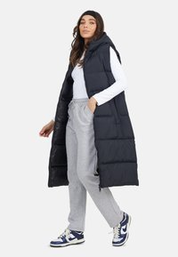 Czarna kamizelka puchowa z kapturem i zapięciem na zamek, z bocznymi kieszeniami i pikowanym wzorem. Nosi się ją na białej koszuli i szarych dresach.