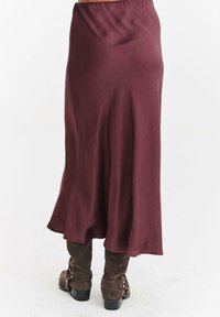 Dlouhá vínová midi sukně nošená přes hnědé semišové kotníkové boty se stříbrnými přezkami na bílém pozadí.
