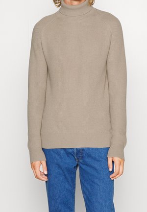Pull à col roulé beige en côtes avec des manches longues et un ourlet ajusté, associé à un jean bleu. Le pull présente un motif de tricot texturé.